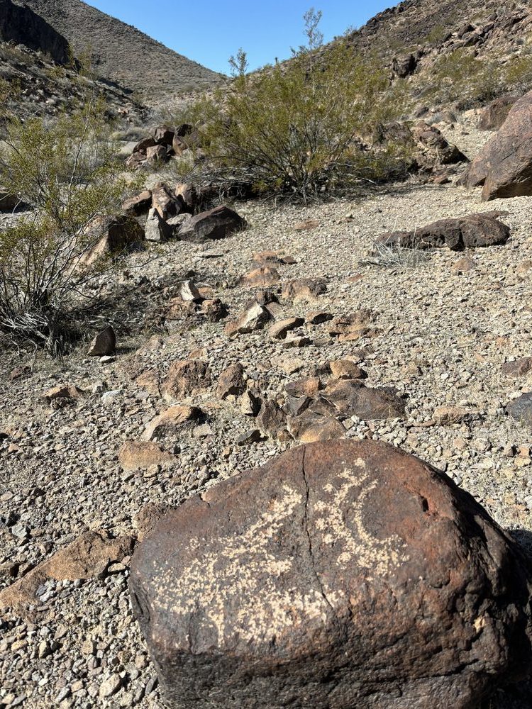 A close-up petroglyph panel carved into a dark desert-varnished boulder in Sloan Canyon, showing a spiral and abstract rock-art design, with a rocky canyon and blue sky in the background