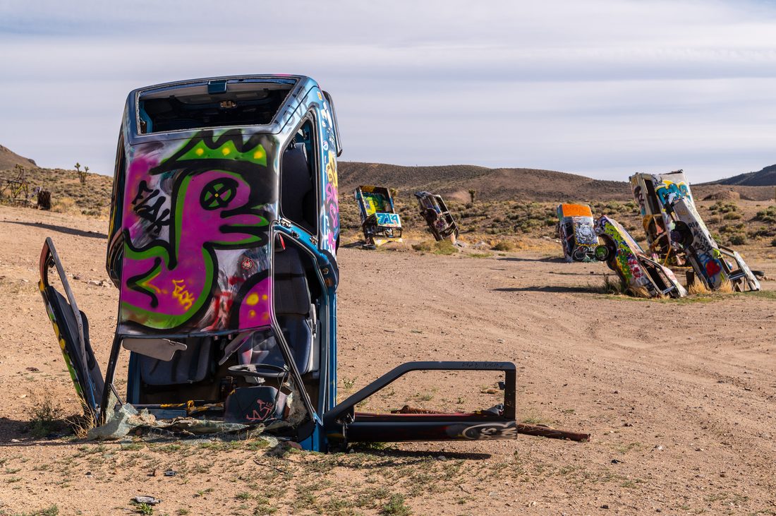 A graffiti-painted van buried nose-down in the desert dirt at the International Car Forest of the Last Church, with several other painted vehicles standing on end in the background near Goldfield, Nevada