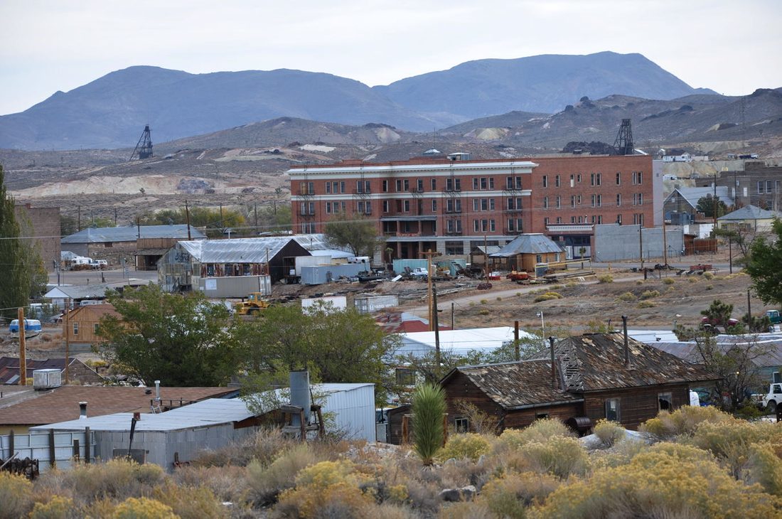 An overview of Goldfield, Nevada showing the vacant multi-story brick Goldfield Hotel amid the town's older buildings, with desert mountains and mine headframes in the background