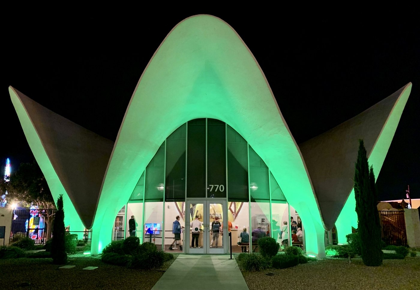 The former La Concha Motel lobby, now the Neon Museum's visitor center, showing its curved shell-shaped Googie-architecture roof lit green at night, with visitors visible inside through the glass entrance
