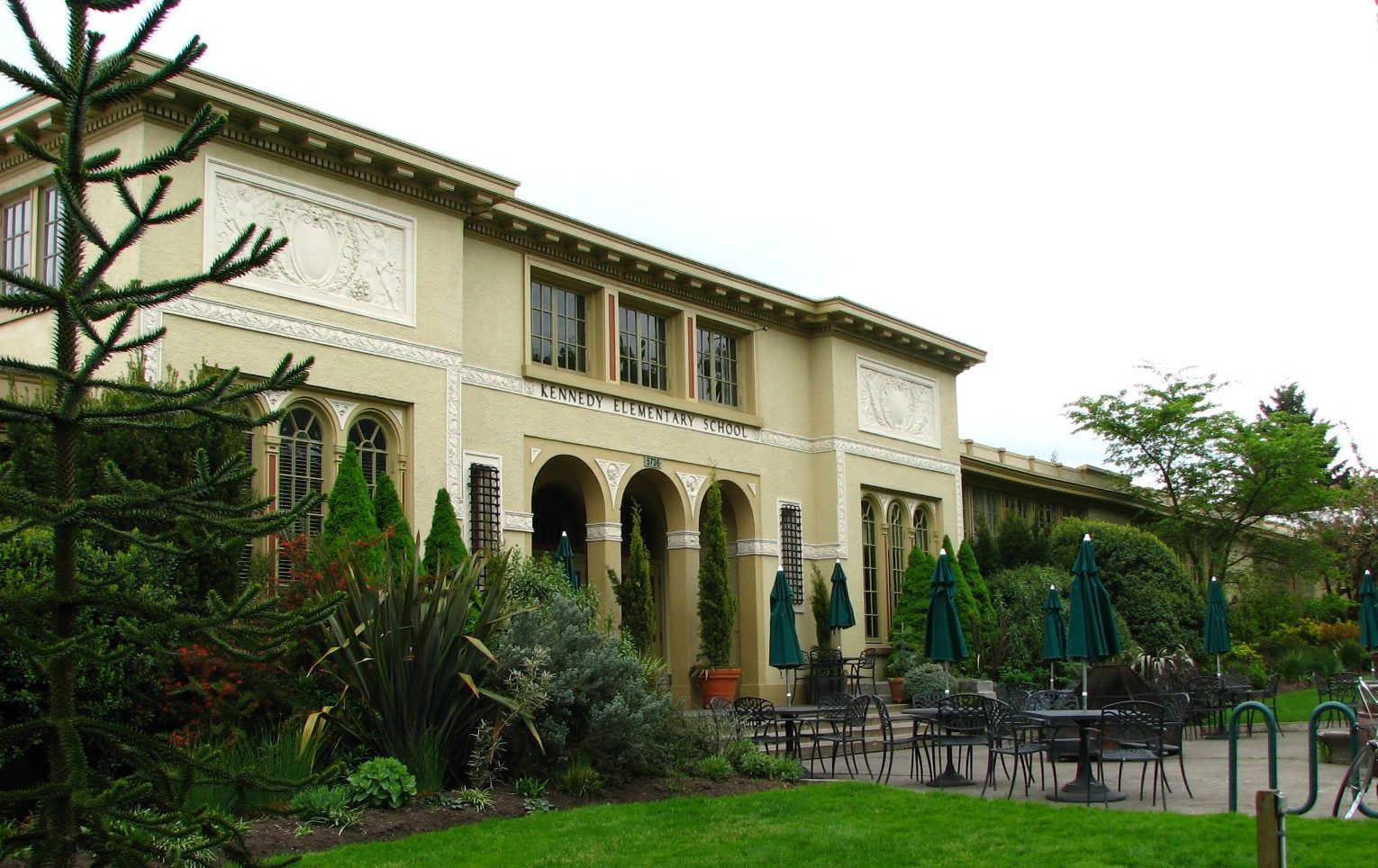 The exterior of McMenamins Kennedy School, a converted 1915 elementary school building in Northeast Portland with a stone entrance, arched windows, and an outdoor patio with green umbrellas