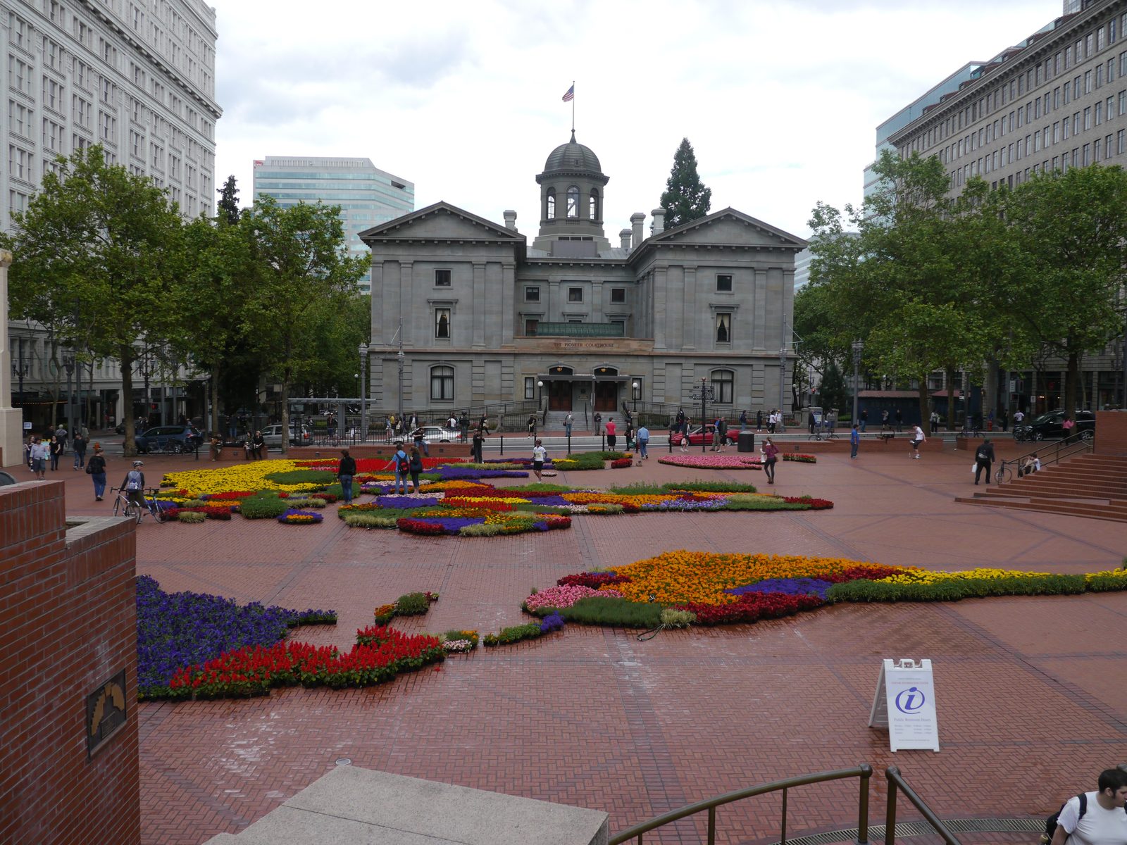 Pioneer Courthouse Square in downtown Portland, showing the historic Pioneer Courthouse building with a domed cupola behind colorful flower-bed displays laid out across the brick plaza