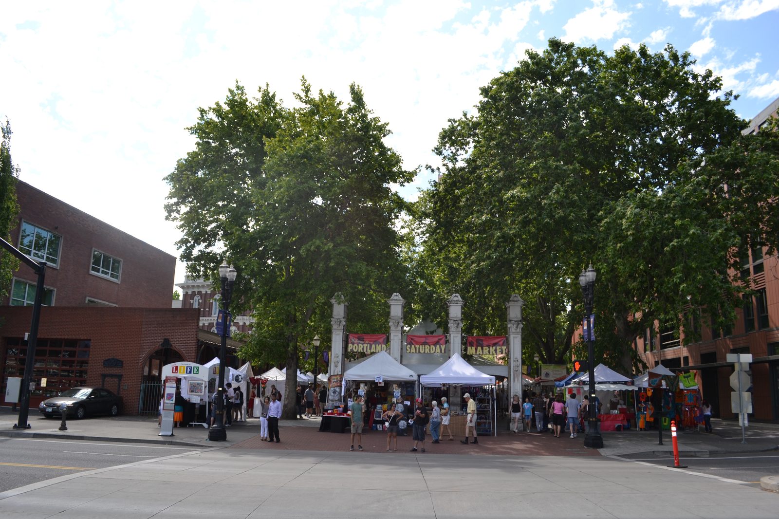 The entrance to Portland Saturday Market, marked by tall stone pillars and a banner reading PORTLAND SATURDAY MARKET, with white vendor tents set up beneath large shade trees