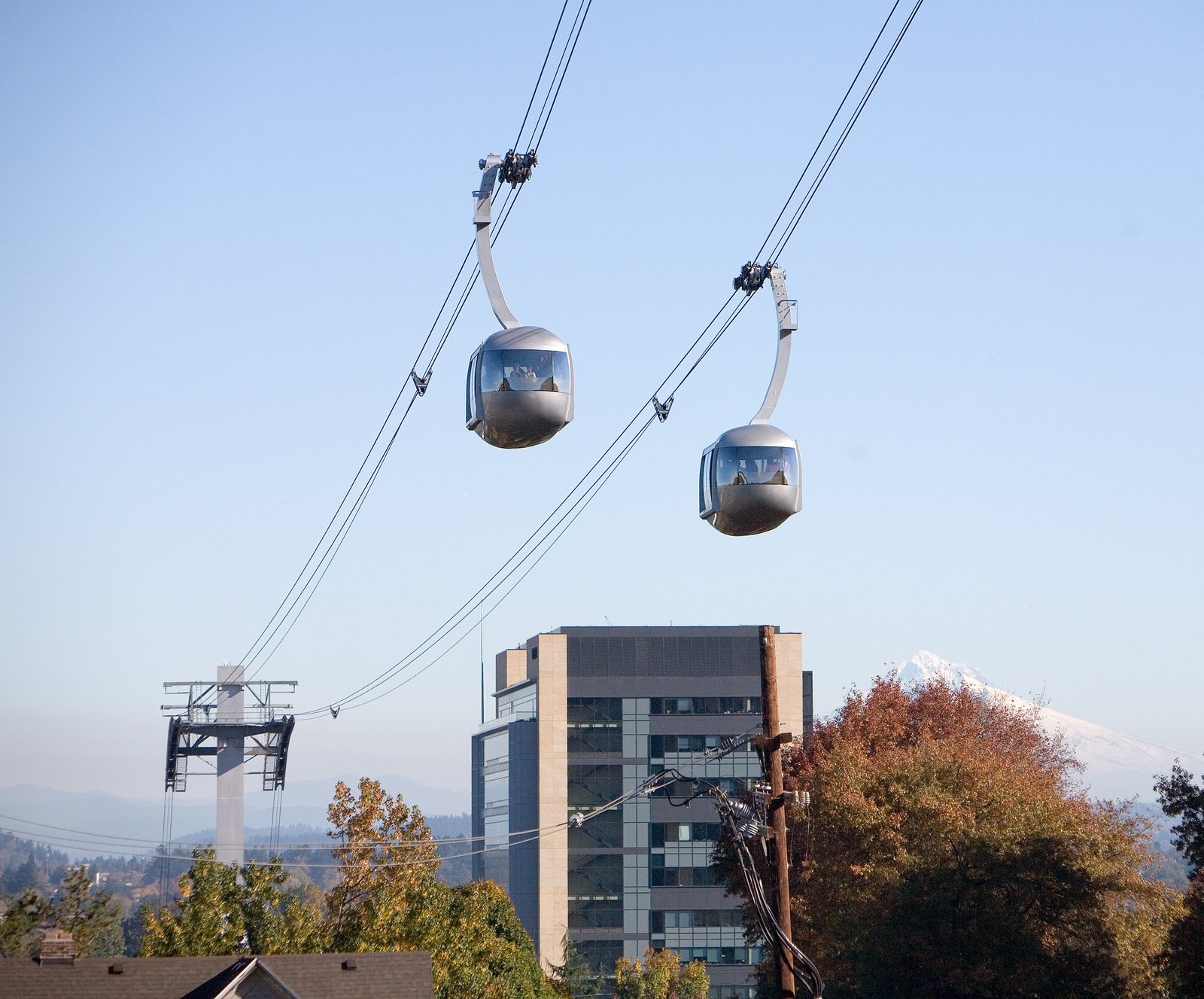 Two silver Portland Aerial Tram cabins suspended on cables above the city, with a hospital tower and snow-capped Mount Hood visible in the background